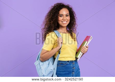 Image of joyful african american student girl with curly hair holding exercise books isolated over violet background