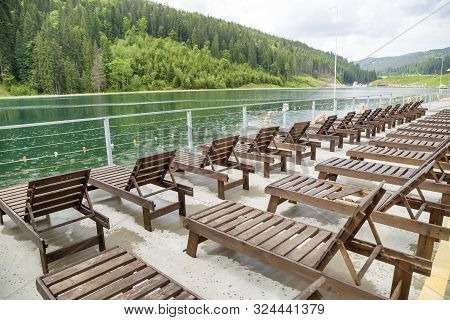 Row Of Empty Wooden Beach Chairs Near Water.