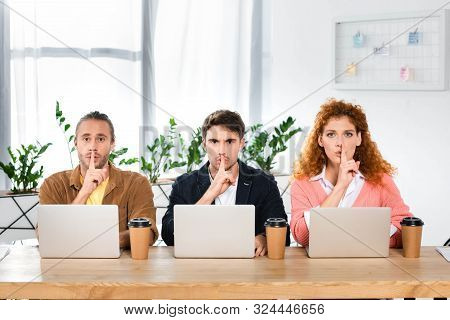 Three Friends Sitting At Table And Showing Shh Gestures In Office