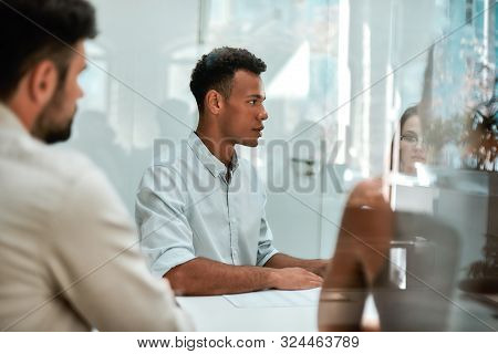 At The Meeting. Young African Man Discussing Something With His Colleagues While Sitting At The Offi