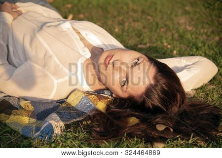A 40-year-old Woman In A White Shirt Lies On A Plaid Blanket, On The Ground, Her Head To The Camera 