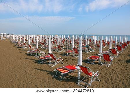 Sun Umbrellas With Deckchairs On The Beach Without People In The Early Morning And The Sea In The Ba