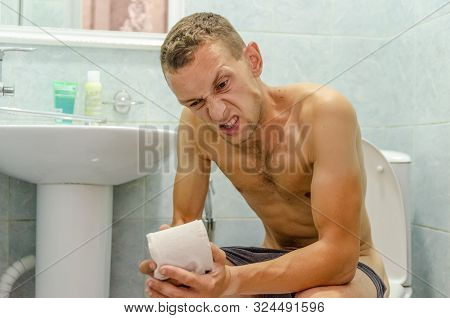 Dark-haired Man In Shorts With Toilet Paper In His Hands Sitting On The Toilet And Writhing In Pain
