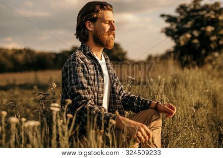 Relaxed Bearded Man With Closed Eyes Sitting In Meadow And Meditating While Establishing Connection 