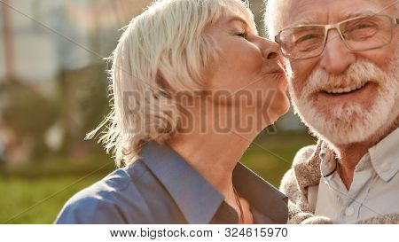 Beautiful Senior Woman Kissing Her Husband On The Cheek While Standing In The Park Together