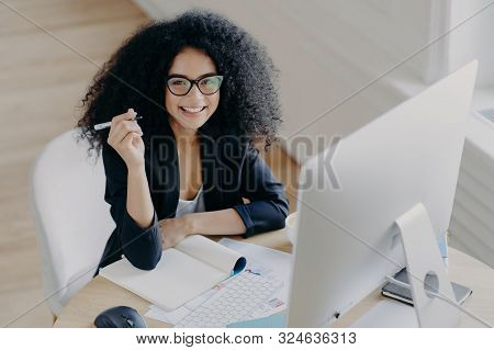 Pleased Dark Skinned African American Woman Holds Pen, Writes Down Some Information, Works As Editor