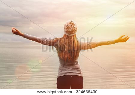 A Beautiful Girl Stands On The Shore With Her Back To The Camera. Hands Up. Sunset. Yoga Classes