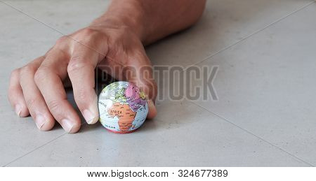 Small Globe Held By Fingers With Grey Concrete Background Seen From The Front. World In Our Hands Wi