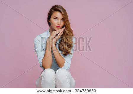 Serious Beautiful Woman With Long Hair, Dressed In Shirt And Trousers, Keeps Palms Pressed Together 