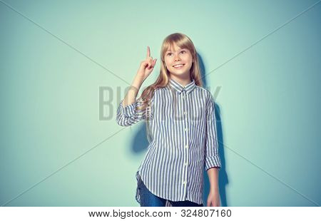 Beauty funny blonde teenage girl has an idea, pointing finger, showing empty copy space for text, blue background. Happy girl presenting point. School girl Proposing product. Advertisement gesture