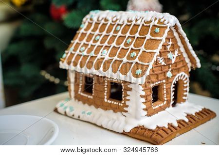 Homemade Christmas Gingerbread House In The Snow. Decorated Christmas Tree In The Background.
