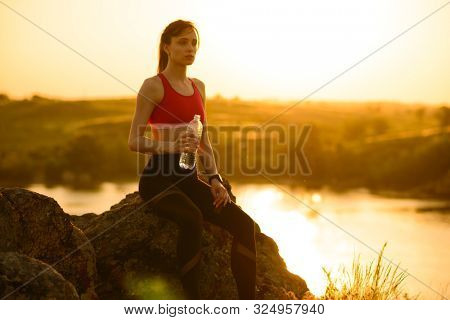 Young Woman Resting and Drinking Water after Trail Running Outdoor. Workout Exercise at Hot Beautiful Summer Sunset. Sport and Healthy Active Lifesyle Concept.