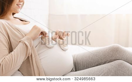 Expecting For Firstborn. Pregnant Woman Playing With Tiny Shoes, Stepping On Her Belly, Empty Space