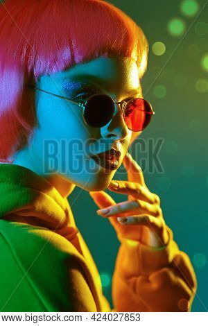 Modern girl with bright makeup and pink hair poses in red sunglasses and yellow hoodie in mixed color light. Sport chic fashion. Youth fashion and beauty trends. 