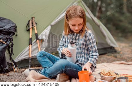 Beautiful preteen girl sitting in the forest in camping and drinking tea from the cup. Cute caucasian child making picnic with tent in the wood