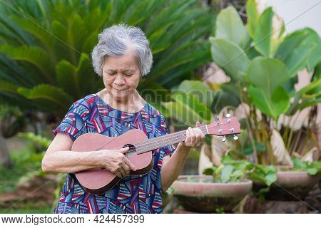 Senior Asian Woman Holding And Looking Ukulele While Standing In A Garden. Relaxing By Singing And P