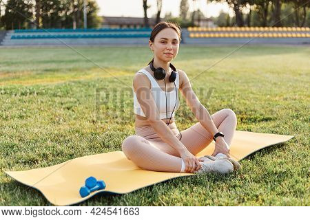 Beautiful Young Female Wearing White Top And Beige Leggins Sitting On Yellow Karemat With Headphones