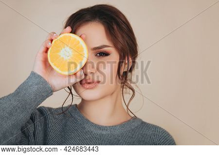 Brunette Female Wearing Wooven Grey Sweater Holding Orange Slice Next To Eye