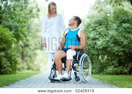 Pretty nurse walking with male patient in a wheelchair in park