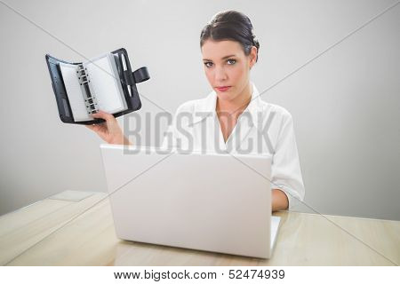 Serious businesswoman working on laptop holding datebook in bright office