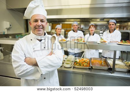 Experienced head chef posing proudly in a modern kitchen with his team in the background