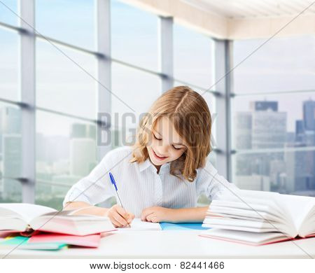 education, people, children and school concept - little student girl sitting at table with books and writing in notebook over classroom background