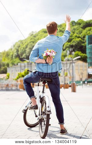 Making a surprise. Handsome young man holding flowers behind his back and waving a hand in sign of salutation while sitting on a bike