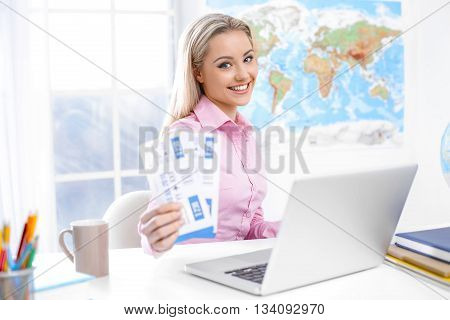 Concept for travel agent. Young blonde woman smiling, looking at camera and showing tickets. Travel agency office interior with big world map and window