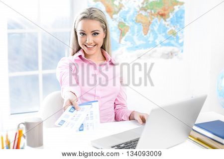 Concept for travel agent. Young blonde woman smiling, looking at camera and showing tickets. Travel agency office interior with big world map and window