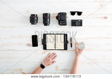 Hands of young man writing in notebook and drinking water on wooden table with blank screen mobile phone, sunglasses, lenses and photo camera