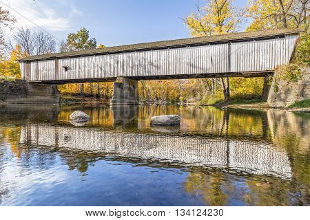 A weathered Darlington Covered Bridge built in 1868 crosses Indiana's Sugar Creek surrounded by colorful fall foliage on a sunny day.