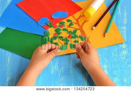 Child tearing colored paper into pieces. Home activity to improve fine motor skill development. Baby play. How to work with paper and glue. Color paper sheets, glue stick, pencils. Blue background