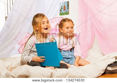 Two little smiling girls with pink wings  reading a book at the bed with net valance