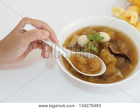 An Asian-forties-lady's hand is using a spoon to scoop Chinese style soup; or braised fish maw in red gravy with eggs, mushroom, and bamboo shoot in white bowl.