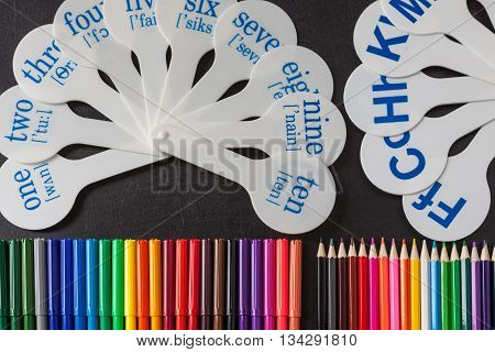 Back to school background with a lot of colorful felt-tip pens and colorful pencils and cards of numerals from one to ten with transcription and consonant letters of alphabet on the black school chalkboard