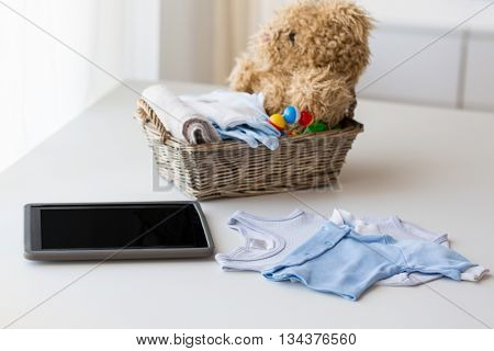 babyhood, motherhood, clothing, technology and object concept - close up of baby clothes and toys for newborn boy in basket with tablet pc computer at home