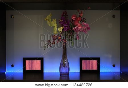 Shelf with lighting decoration flowers in bowl and frames against multicolor backlit plastic panel