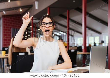 Cheerful excited young businesswoman in glasses pointing up and having an idea working in office