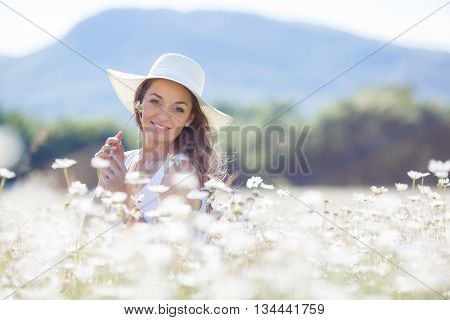 Portrait of beautiful young woman in a white hat with a wide brim, brunette with long curly hair, in a white summer dress,a beautiful smile, on a neck a gold chain, aposing in a white field of blooming daisies in a mountain area