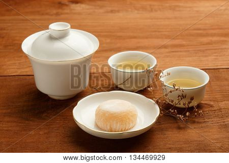 Gaiwan and cups of white chinese porcelain with green tea standing on wooden table with delicious japanese mochi rice cake on white plate decorated with dried flower. Shallow dof focus on foreground.