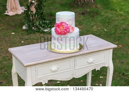 Vintage wooden table with the wedding cake standing on a green lawn. Cake decorated with fresh peonies