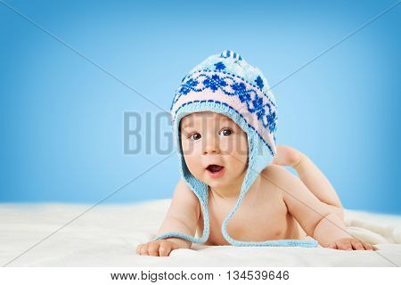 Eight month old baby in knitted winter hat lying on soft blanket with blue background