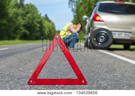 Sad man after car breakdown broken car and spare wheel on the roadside. Focus on red triangle warning sign
