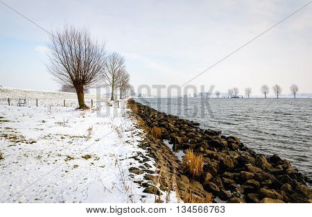 Wide Dutch river in the winter season. A layer of white snow has fallen on the stones on the bank of the river.