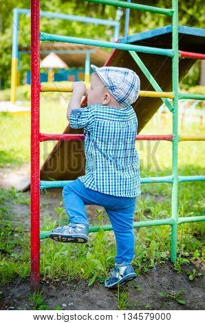 Little toddler climbing the ladder having fun at playground in summer. Cute young boy kid or child playing outdoors on playground. Stylish little baby boy in blue shirt pants and hat. Summer time.