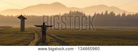 View from behind of a man in winter jacket and hat standing on country road in the middle of beautiful landscape with hayrack on his left with his arms outstretched at dusk.