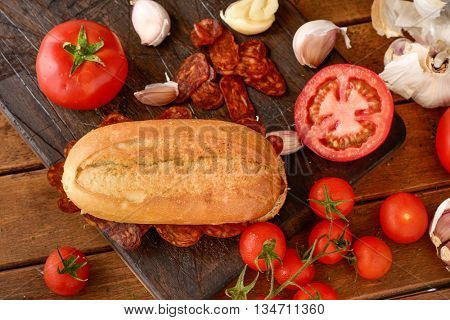 high-angle shot of a spanish bocadillo de chorizo, a sandwich made with sliced chorizo, a pork sausage typical of Spain, on a rustic wooden table with some tomatoes and garlic cloves