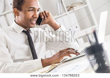 Side view of friendly african american businessman using notepad and laptop on office desktop while having mobile phone conversation. Bookshelf with documents in the background