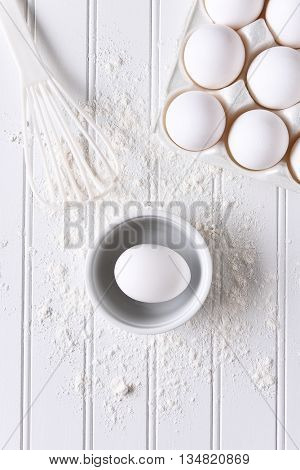 White on White baking still life. Top view of items for baking: eggs, flour and a whisk on a white beadboard surface. 
