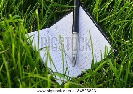 White note-book with black pen and empty white pages on green grass field